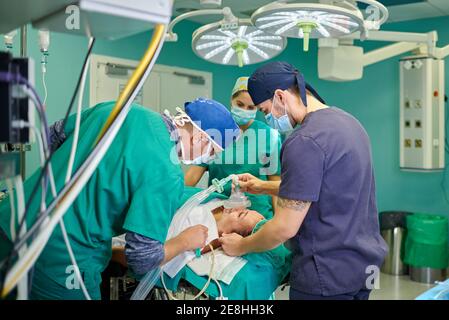 Side view of anonymous male anesthesiologist in medical uniform and cap holding anesthesia mask on face of unrecognizable patient lying on couch in op Stock Photo