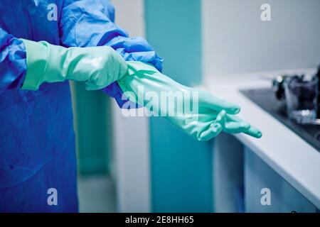 Crop anonymous medical worker in surgical gown putting on rubber gloves before disinfecting endoscopy instruments in cleaning workstation Stock Photo