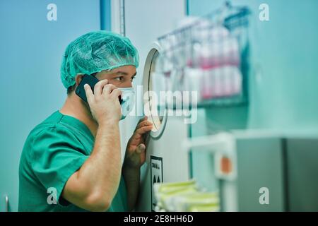 Side view of concentrated young male doctor in uniform and mask having phone conversation and looking through round window of operating room door duri Stock Photo