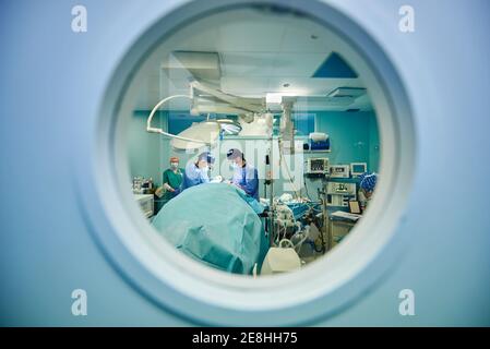 Through round window of unrecognizable doctors in uniforms and masks performing surgery in modern operating room Stock Photo