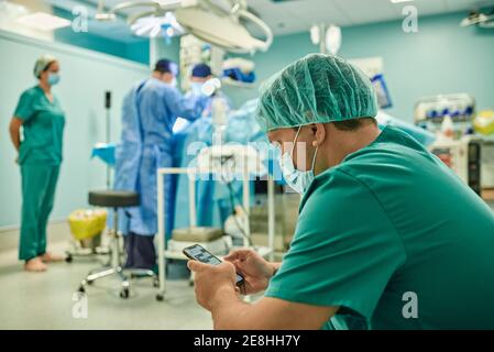 Side view of anonymous concentrated young male assistant in medical uniform and mask messaging on mobile phone while team of professional surgeons per Stock Photo
