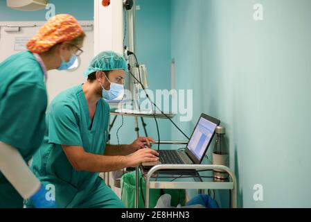 Side view of anonymous young female and male assistants in scrubs and masks using laptop during medical procedure in operating room Stock Photo