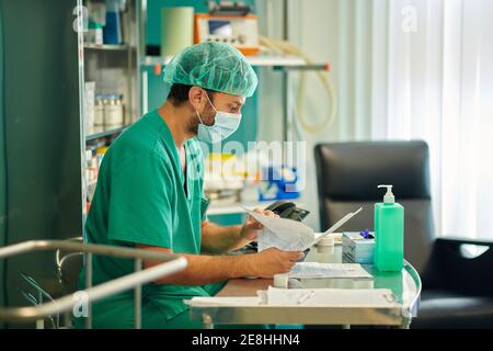 Side view of young concentrated male doctor in medical uniform and mask checking test results while sitting at table in hospital cabinet Stock Photo