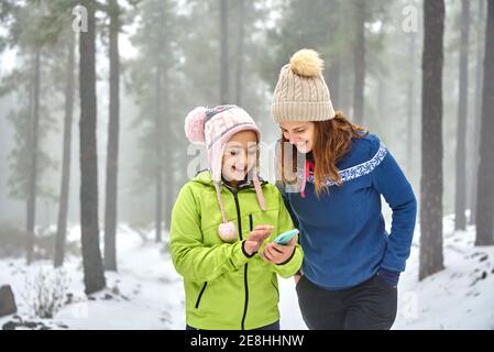 Happy smiling mother and daughter in outerwear browsing mobile phone while walking together in snowy frozen woodland Stock Photo