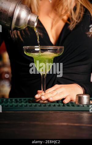 Crop faceless female bartender in stylish outfit pouring alcohol cocktail from shaker into elegant margarita glass in restaurant Stock Photo