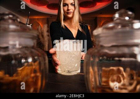 Content young female bartender with long blond hair serving cold alcohol cocktail in stylish restaurant looking at camera Stock Photo