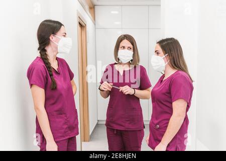 Group of young content nurses in medical uniform and masks having conversation in modern hospital corridor Stock Photo