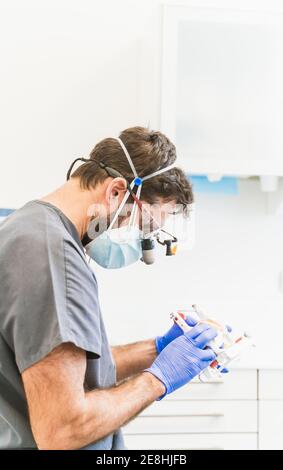 Side view focused male dental technician in medical uniform and binocular eye loupe glasses working with dental prosthetics in modern clinic Stock Photo