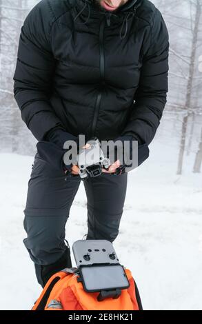 Crop young male wearing black warm outwear holding modern folded drone while standing in snowy freezing woodland Stock Photo
