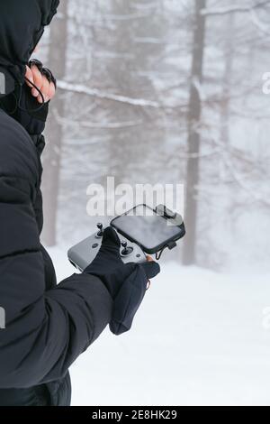 Cropped unrecognizable male wearing warm black jacket with hood standing on snowy terrain with drone remote control Stock Photo