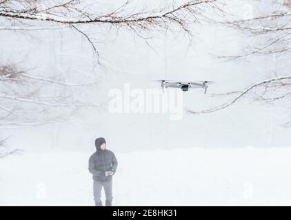Male wearing warm winter clothes holding remote control of modern UAV flying near in frozen woodland Stock Photo