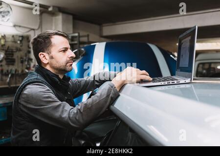 Side view of young focused bearded master watching netbook screen on top of vehicle in auto workshop Stock Photo