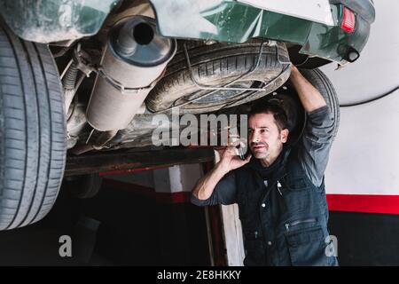 Young focused male auto master speaking on cellphone while checking up rear part of transport and looking away in workshop Stock Photo