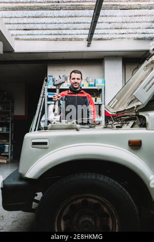 Young content bearded maintenance man showing metal instrument while looking at camera near transport in auto workshop Stock Photo