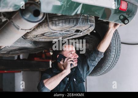 Young focused male auto master speaking on cellphone while checking up rear part of transport and looking up in workshop Stock Photo