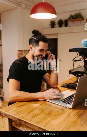 Smile barista sitting at wooden counter with coffee machine and working while talking on smartphone and using laptop Stock Photo