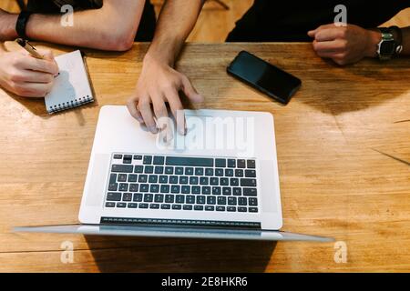 From above of crop male baristas sitting at wooden counter in coffee shop and working together while using laptop and taking notes in notebook Stock Photo