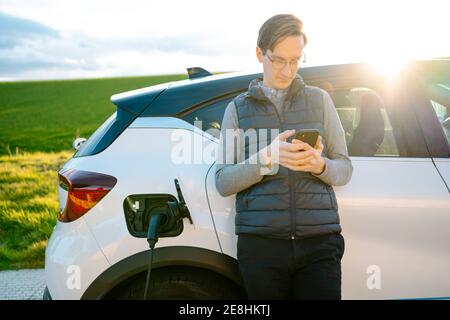 Concentrated young male driver browsing mobile phone while leaning on modern hatchback car parked near green field during refueling in sunlight Stock Photo