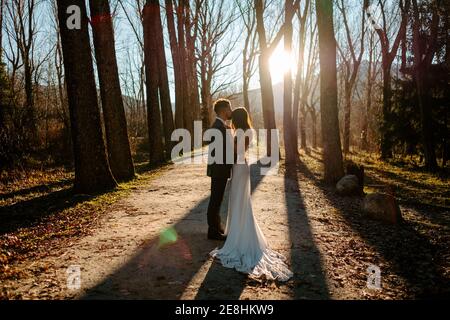 Side view of unrecognizable young groom in elegant suit kissing forehead of graceful bride in white dress while standing in autumn forest on wedding d Stock Photo