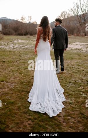 unrecognizable Full length of stylish young ethnic bride and groom holding hands and looking at camera while standing in autumn forest near leafless t Stock Photo