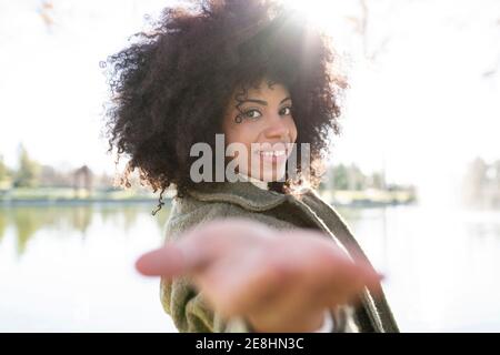 Side view of positive young black lady with Afro hairstyle in trendy outfit reaching out hand towards camera and smiling while resting near lake in au Stock Photo