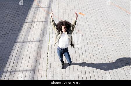 From above of expressive young black lady with Afro hairstyle in stylish clothes standing on paves street with raised arms and smiling on sunny day Stock Photo