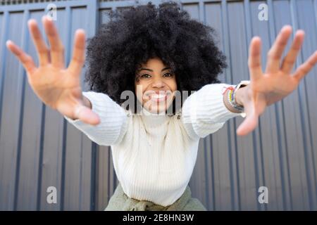 Cheerful young African American lady with curly hair in casual clothes showing two hands and looking at camera on street Stock Photo