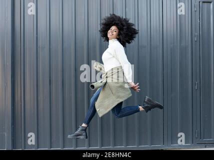 Side view of happy young African American Woman with curly hair in trendy outfit smiling and looking at camera while jumping on street Stock Photo
