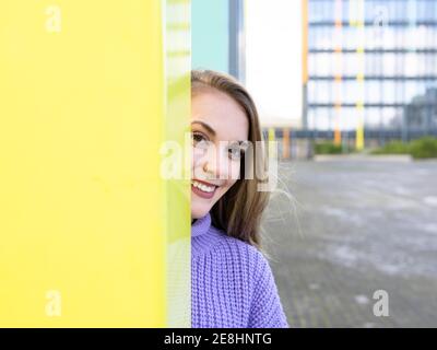 Attractive young female in blue sweater standing behind yellow wall on street and looking at camera with charming smile Stock Photo