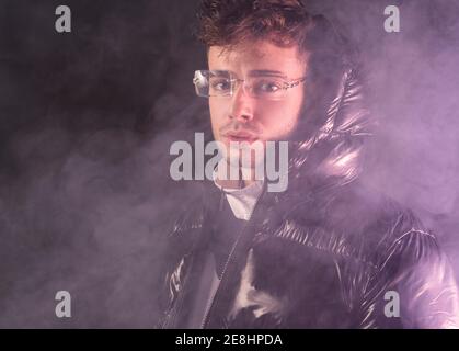 Thoughtful young unshaven guy in trendy outfit against black background and smoke Stock Photo