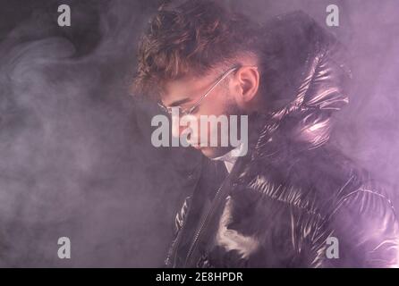 Thoughtful young unshaven guy in trendy outfit against black background and smoke Stock Photo