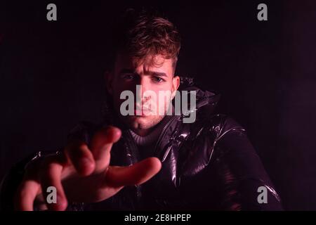 Thoughtful young unshaven guy in trendy outfit reaching out hand towards camera against black background Stock Photo