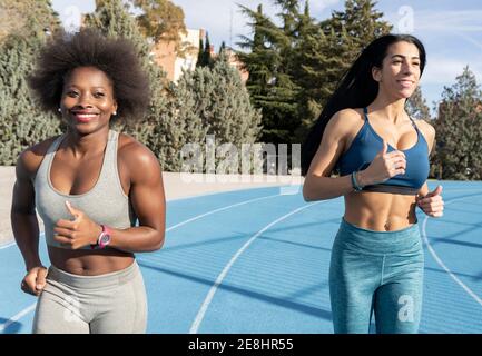 Multiracial female runners in sportswear running together at stadium while smiling and enjoying workout on sunny day Stock Photo
