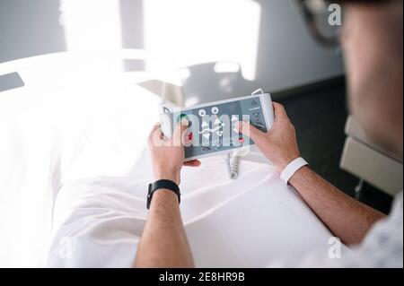 Adult male wearing patient gown and eyeglasses browsing hospital bed remote control on bed in light ward in modern hospital Stock Photo