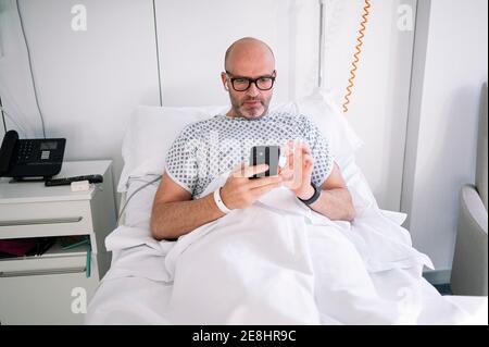 Concentrated adult male wearing patient gown and eyeglasses browsing phone on bed in light ward in modern hospital Stock Photo