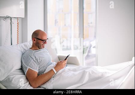 Positive adult male wearing patient gown and eyeglasses browsing phone on bed in light ward in modern hospital Stock Photo