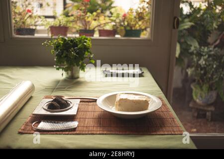 Tasty soft cheese in plate near truffles and grater on straw mat at home with potted plants Stock Photo