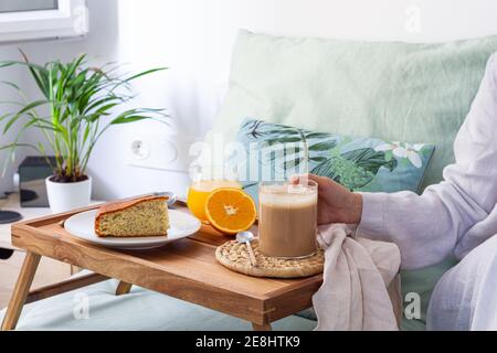 Unrecognizable crop female sitting on bed with cup of coffee and tray served with sponge cake and glass of juice while having breakfast at home Stock Photo
