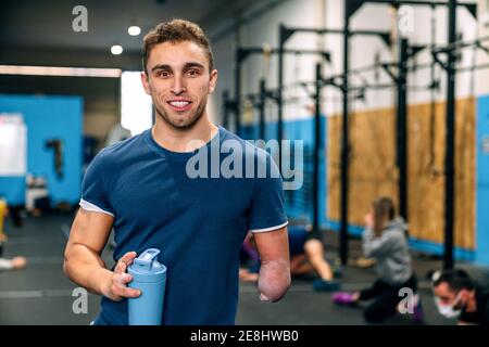 Young disabled happy male athlete in sportswear holding bottle of water while looking at camera during workout near unrecognizable partners in gymnasi Stock Photo
