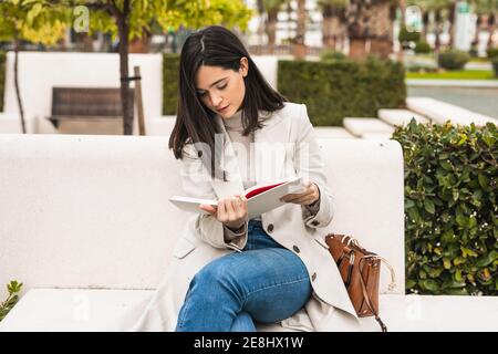 Female entrepreneur sitting on bench in park and checking schedule in notepad Stock Photo