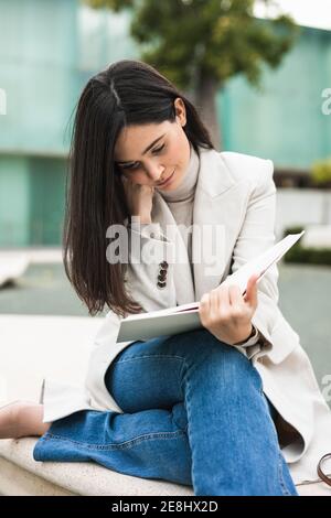 Pensive female entrepreneur sitting on bench and leaning on hand while reading book in city Stock Photo