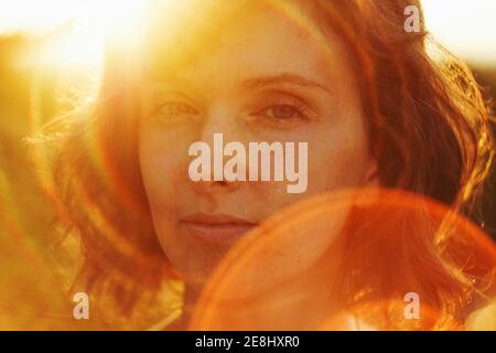 Crop thoughtful adult female resting in nature and looking at camera pensively under bright sunlight during golden hour Stock Photo