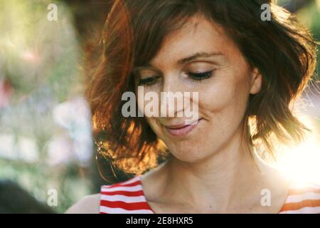 Crop thoughtful happy adult female resting in nature and looking down pensively under bright sunlight during golden hour Stock Photo