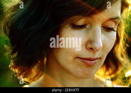 Crop thoughtful adult female resting in nature and looking down pensively under bright sunlight during golden hour Stock Photo