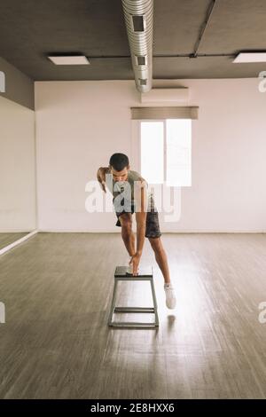 Full length of focused young muscular sportsman practicing Squat Pop Over exercise on step platform while exercising alone in light studio Stock Photo
