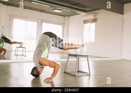 Side view of young muscular male gymnast doing Pike Push Ups with legs on step platform during training in studio near mirror Stock Photo