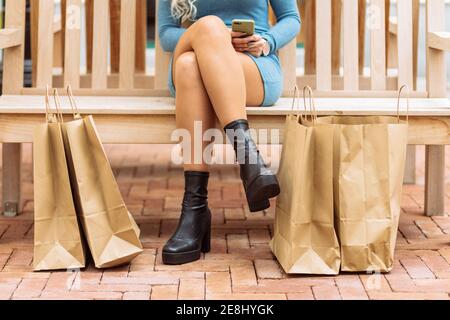 Crop unrecognizable female sitting on bench near paper bags and browsing smartphone after shopping in city Stock Photo