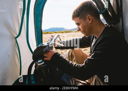 Side view of young dreamy male traveler in casual outfit resting in camping tent and checking his backpack on sunny day Stock Photo