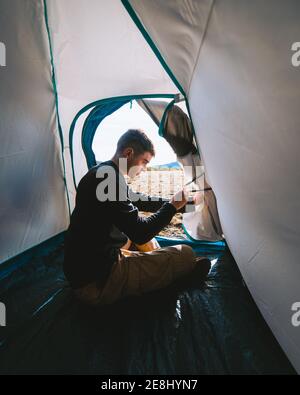 Side view of concentrated young male camper in casual outfit opening tent door while recreating during hiking trip on sunny day in mountains Stock Photo