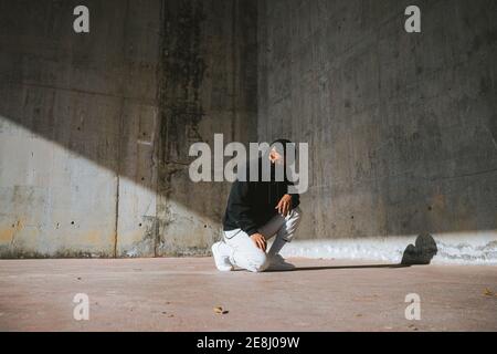 Unrecognizable young male in hoodie and mask sitting on street near concrete wall Stock Photo